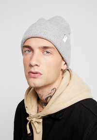 Grey ribbed beanie made of soft knit material, featuring a small white label. Worn with a beige hoodie and black jacket.