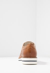 Scarpa in pelle marrone con dettagli perforati, punta rotonda e suola bianca con striscia blu. Vista posteriore che enfatizza texture e design.