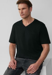 Czarna, krótka koszulka z dekoltem w serek, wykonana z miękkiego materiału, z subtelnym logo na piersi. Model w połączeniu z szarymi dżinsami, siedzący na jasnej powierzchni.