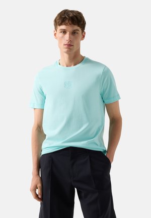 Młody mężczyzna w jasnoniebieskiej koszulce z krótkim rękawem z małym logo na piersi i czarnych spodniach, stojący na jednolitym tle.