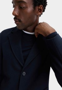 Marinblå blazer i strukturerat tyg med krage med notch, enkelknappsförslutning och smal passform. Bärs över en mörk topp med rund halsringning.