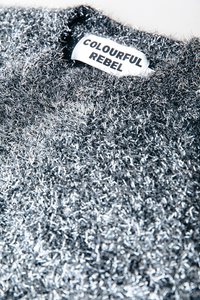 Detailný záber na striebornú kovovú textúru látky s bielou etikettou na oblečení, na ktorej sú tučne napísané slová "COLOURFUL REBEL" v čiernych písmenách.