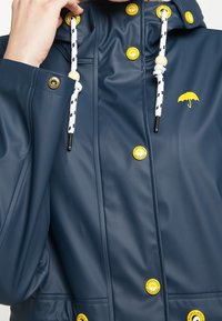 Imperméable marine avec boutons jaunes, cordons blancs avec accents noirs, perles en bois et logo parapluie jaune sur la poitrine.