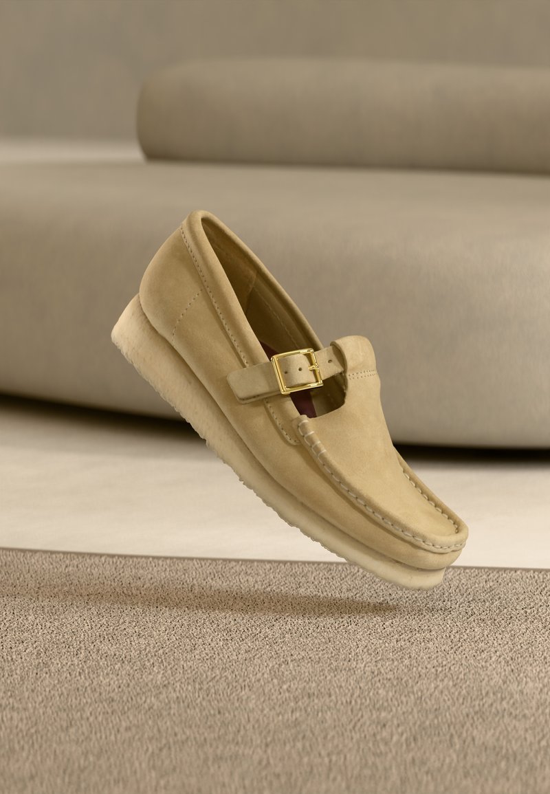 Beiger Wildleder-Slipper mit einem goldenen Schnallenriemen, detaillierter Naht und einer Krepsohle, präsentiert auf einem strukturierten Teppich mit beigem Sofa im Hintergrund.