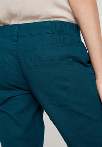 Tealfärgade bomullsbukser med elastisk midja, med två bakfickor och stickad detalj, som visar en jämn, lätt textur.