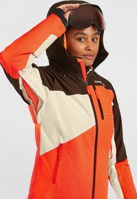Veste de ski avec un design bicolore en orange, marron et crème, avec des poches zippées et une capuche. Fabriquée en matériau durable et résistant aux intempéries.