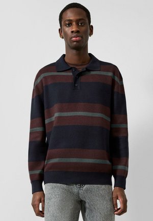 Jeune homme portant un polo à manches longues avec de larges rayures horizontales bleu marine, marron et grises, ainsi qu’un jean gris clair, se tenant devant un fond uni.