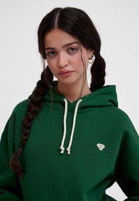 Zielona bluza z kapturem z sznurkiem, z małym haftowanym logo na klatce piersiowej. Materiał wydaje się miękki, o luźnym kroju i ciekawej teksturze.