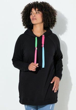 Mujer de cabello rizado vistiendo una sudadera negra con cordones multicolores y jeans azules desgastados, de pie contra una pared blanca.