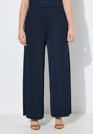 Vrouw die marineblauwe wijde broek draagt gecombineerd met bruine sandalen met open teen, gefotografeerd van taille tot voeten tegen een witte achtergrond.