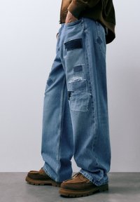 Wijde spijkerbroek in lichtblauw met diverse patches en verweerde details. Kenmerken een losse pasvorm, meerdere stoftexturen en een rafelige zoom.