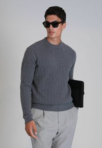 Grauer Kabelstrickpullover mit rundem Ausschnitt und gerippten Bündchen, kombiniert mit hellgrauen Hosen und einer schwarzen Clutch. Sonnenbrillen runden den Look ab.