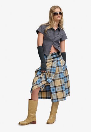 Jeune femme portant des lunettes de soleil, un t-shirt gris à manches courtes, une jupe à carreaux bleue et beige, des chauffe-bras noirs et des bottes hautes beige.