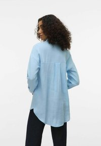 Light blue long-sleeve shirt in textured fabric, featuring a back pleat and curved hem. Paired with black trousers.