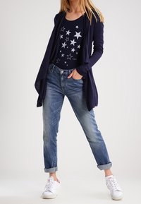 Marinblå cardigan över en marinblå topp med stjärnmönster, tillsammans med ljusblå jeans med uppvikta ärmar och vita sneakers med röda accenter.