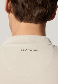 Koszulka polo w jasnym beżowym materiale o teksturowanym splotcie. Posiada prążkowany kołnierzyk oraz brązowe haftowane logo z tyłu.