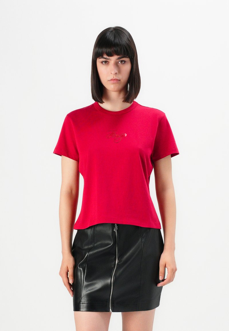 Donna con capelli lunghi e scuri, che indossa una maglietta rossa a maniche corte e una mini gonna di pelle nera con cerniera frontale, in piedi su uno sfondo bianco.