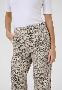 Pantalons beiges avec un motif léopard noir. Ils présentent une coupe classique, des poches avant et une fermeture à bouton à la taille.