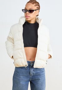 Puffjakke i lys beige med quiltet design, høy krage og glidelåslukkinger på sidene. Brukt med en ribbestrikket svart topp og blå jeans.