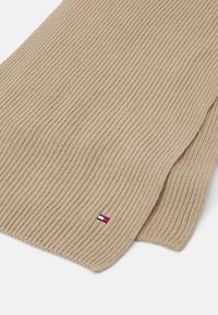Beige ribbet strikket tørklæde med et lille rødt, hvidt og blåt logo. Teksturen er blød og strækbar, med rene, færdige kanter.
