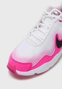 Nike sportcipő fehér hálós felsőrésszel, rózsaszín átmeneti részletekkel és fekete logóval. Vastag, textúrált talppal rendelkezik, és fűzős elülső résszel.