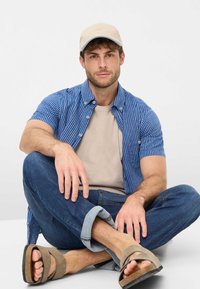 Camicia a maniche corte a righe blu sopra una t-shirt beige, abbinata a jeans scuri arrotolati e sandali marroni senza lacci. Cappellino chiaro in testa.