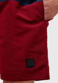 Røde stoffshorts med en jevn tekstur, med en svart firkantet lapp med teksten "JEANS CODE" nær kanten. Marineblå aksent i midjen.