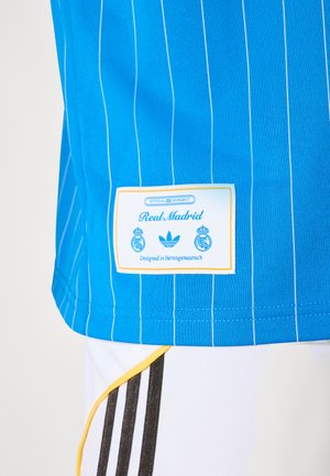 Modrý prúžkovaný dres Realu Madrid s oficiálnou značkou a biele šortky s čiernymi pruhmi a žltým lemovaním.
