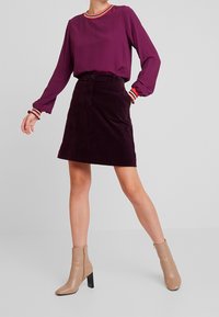 Blusa viola a maniche lunghe con colletto a costine multicolore, abbinata a una gonna in velluto a coste di un viola scuro e stivaletti beige con tacco nero.