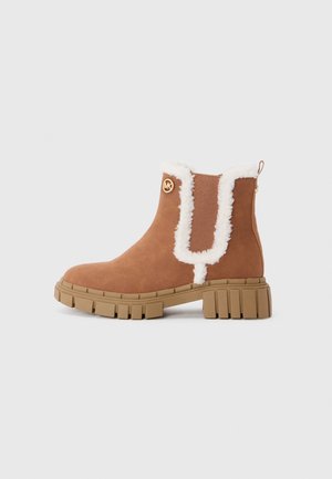 Bottine en suede tan avec une finition en fausse fourrure blanche, semelle en caoutchouc épaisse, panneau élastique sur le côté et détail doré du logo. Comprend une languette à l'arrière.