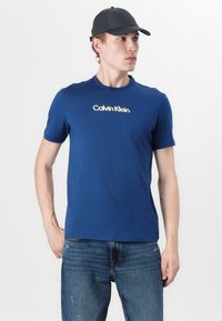 Ναυτικό μπλε βαμβακερό μπλουζάκι με στρογγυλή λαιμόκοψη και κοντά μανίκια, με λευκό λογότυπο Calvin Klein στο στήθος. Φορεμένο με μπλε τζιν.