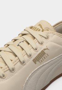 Μπεζ αθλητικό παπούτσι Puma Tackle με μπεζ πλεγμένα κορδόνια, ανάγλυφη λωρίδα στο πλάι και λογότυπα Puma στη γλώσσα και στο πλευρό.