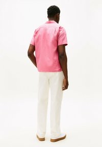 Camicia rosa a maniche corte con una texture liscia, abbinata a pantaloni bianchi e sneaker bianche. Cucitura sul retro e dettaglio del colletto ben visibili.