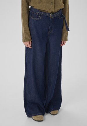 Person, die dunkelblaue Jeans mit weitem Bein und passendem Gürtel, ein beigefarbenes Langarm-Strickoberteil und beige Schuhe trägt, steht vor hellem Hintergrund.