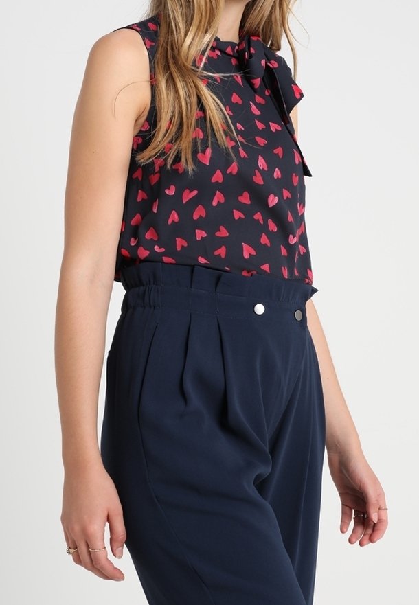 Mouwloze blouse met een marineblauwe achtergrond en een rood hartpatroon, met een strikdetail bij de hals. Hoge taille marineblauwe broek met plooien.