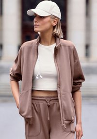 Brown zip-up jacket with side pockets, paired with a beige crop top and matching drawstring pants. A light-colored cap completes the look.