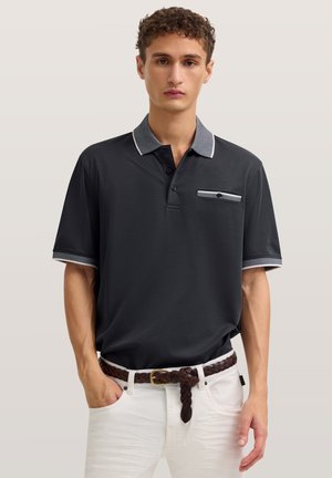 Junger Mann trägt ein schwarzes Polohemd mit grauem Kragen und Taschenapplikation, weiße Hose und einen braunen geflochtenen Gürtel, steht mit einer Hand in der Tasche.