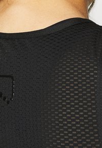 Top deportivo negro con tejido texturizado similar a malla; presenta un escote redondeado y un sutil detalle de logo en la parte posterior.