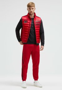 Rote gepolsterte Weste mit hohem Kragen und schwarzen Text-Logos. Getragen über einem schwarzen Sweatshirt und roten Hosen mit seitlichen Streifen, abgerundet mit weißen Sneakern.