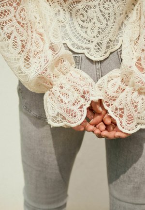 Blondebluse med indviklede hvide blomsterdekorationer og bølgede manchetter, kombineret med tætsiddende lysegrå jeans. Hænder er synlige forneden.