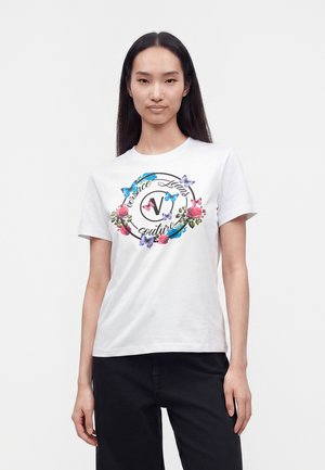 Γυναίκα που φοράει λευκό T-shirt με floral και πεταλούδα σχέδιο γύρω από το κυκλικό λογότυπο Versace Jeans Couture, σε συνδυασμό με μαύρο παντελόνι.