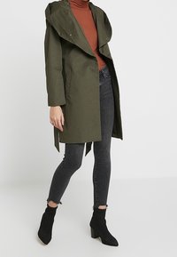 Manteau trench vert olive avec un grand col, des boutons sur le devant et des poches latérales. Porté sur un col roulé marron, assorti à un jean skinny gris et des bottines noires.