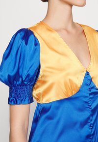Robe en satin présentant un corsage bleu avec des manches bouffantes et un col jaune. Elle a un décolleté en V profond et une fermeture par boutons.