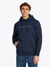 Jonge man in een marineblauwe Tommy Jeans hoodie met een voorzak en jeans, staand tegen een effen lichte achtergrond.