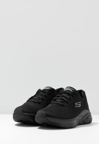 Zapatillas deportivas negras con parte superior de malla, diseño con cordones, cuello acolchado, logotipo en el lateral y suela texturizada para un mejor agarre.