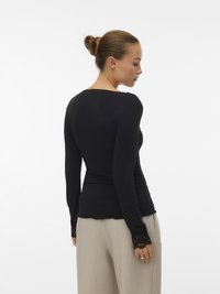 Žena s punđom, odjevena u crnu majicu dugih rukava s čipkastim manžetama i bež širokim hlačama, okrenuta u stranu na bijeloj pozadini.