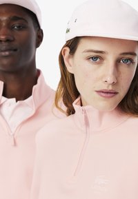 Felpa rosa chiaro con zip e colletto alto, logo Lacoste. Indossata con un cappellino rosa, texture morbida, vestibilità casual e dettagli minimi.