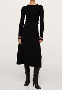 T-shirt noir à manches longues en côtes avec des poignets rayés rouges et blancs, associé à une jupe midi noire. Des bottes noires à talons aiguilles hauteur genou complètent le look.