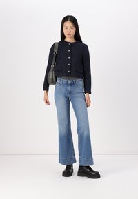 Mornarska teksturirana skraćena jakna sa srebrnim dugmadi, uparena sa svijetloplavim zvonastim farmerkama i crnim platformskim čizmama. Model drži sivi crossbody ranac.