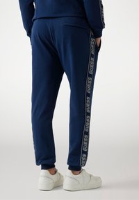 Navy blue joggers made from soft fabric, featuring a side stripe with "GUESS" logo. Includes a back pocket and ribbed cuffs.